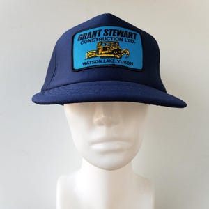 May include: A navy blue trucker cap with a rectangular patch displaying the text "GRANT STEWART CONSTRUCTION LTD." and a yellow bulldozer graphic. The patch also includes "WATSON LAKE, YUKON."
