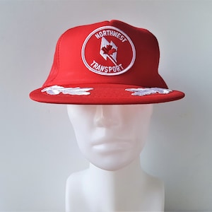 Può includere: Cappellino da camionista rosso con retro in rete bianca e visiera curva. Il cappello presenta una toppa circolare con le parole "NORTHWEST TRANSPORT" e un design a foglia d'acero. Disegni bianchi a forma di nuvola sono ricamati sulla visiera.