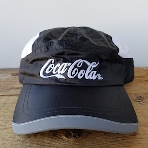 Puede incluir: Gorra de running negra con un logotipo de Coca-Cola blanco bordado en la parte delantera. La gorra tiene una visera gris y una parte trasera de malla.