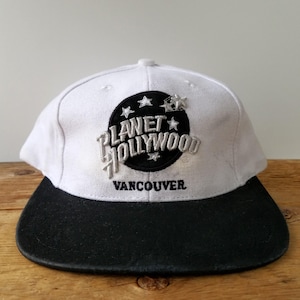 May include: A white and black Planet Hollywood Vancouver baseball cap. The cap features the Planet Hollywood logo in a black circle with silver text and stars. The brim is black.