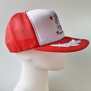 May include: Red and white trucker hat with a mesh back. The front panel is white with a graphic and text. The brim is red with a white cloud design. The hat has the text "12th Annual Winter Games All-Stars".