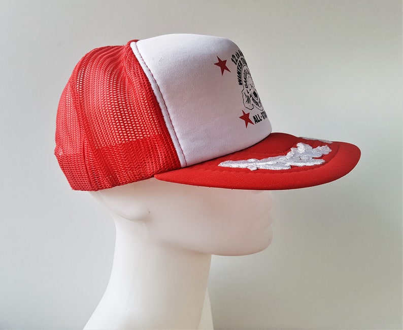 May include: Red and white trucker hat with a mesh back. The front panel is white with a graphic and text. The brim is red with a white cloud design. The hat has the text "12th Annual Winter Games All-Stars".