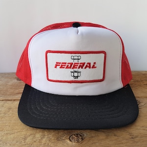 May include: A trucker hat with a red mesh back and a black brim. The front panel is white with a rectangular patch featuring the word "FEDERAL" in red, along with a graphic of a bolt and nut.