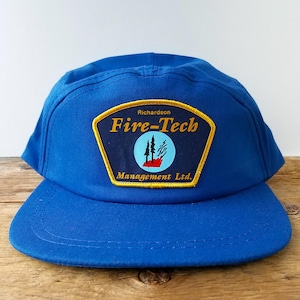 May include: A blue baseball cap with a flat brim. The cap features a patch with the words "Fire-Tech" and "Management Ltd." in gold lettering. The patch also includes a graphic of trees and a red flame.