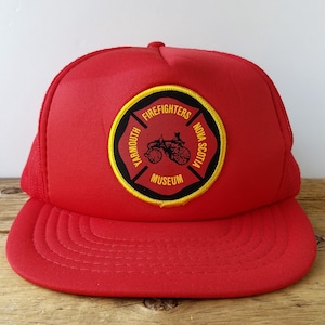 Op de afbeelding: Een felrode truckerpet met een ronde patch aan de voorzijde. De patch heeft een zwart midden met een gele rand en toont de tekst YARMOUTH FIREFIGHTERS MUSEUM NOVA SCOTIA rondom een afbeelding van een antieke brandweerwagen.