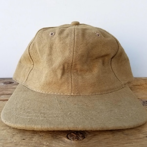 May include: A tan baseball cap with a curved peak. The cap is made of a textured fabric and has a button on top. The cap is on a wooden surface.