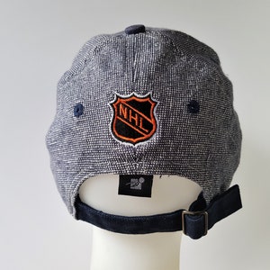 May include: A navy blue baseball cap with a textured pattern. The cap features the NHL logo in orange, black, and white on the back. The adjustable strap and button are navy blue. The Starter logo is visible.