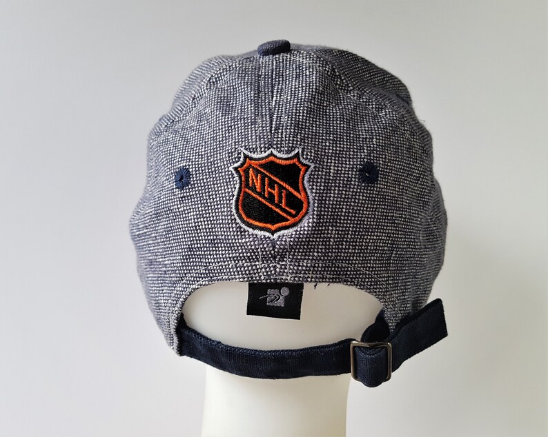 May include: A navy blue baseball cap with a textured pattern. The cap features the NHL logo in orange, black, and white on the back. The adjustable strap and button are navy blue. The Starter logo is visible.