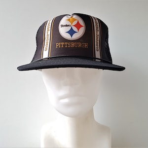 Peut inclure: Casquette de camionneur Pittsburgh Steelers noire et dorée. La casquette présente le logo des Steelers et le mot "PITTSBURGH" sur le devant. La casquette a un dos en filet et une visière incurvée.