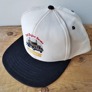 Op de afbeelding: Een wit en zwarte baseball cap met de tekst "Truck Engine Service FINNING" en een afbeelding van een vrachtwagen. De cap heeft een zwarte klep en een zwarte knop bovenop.