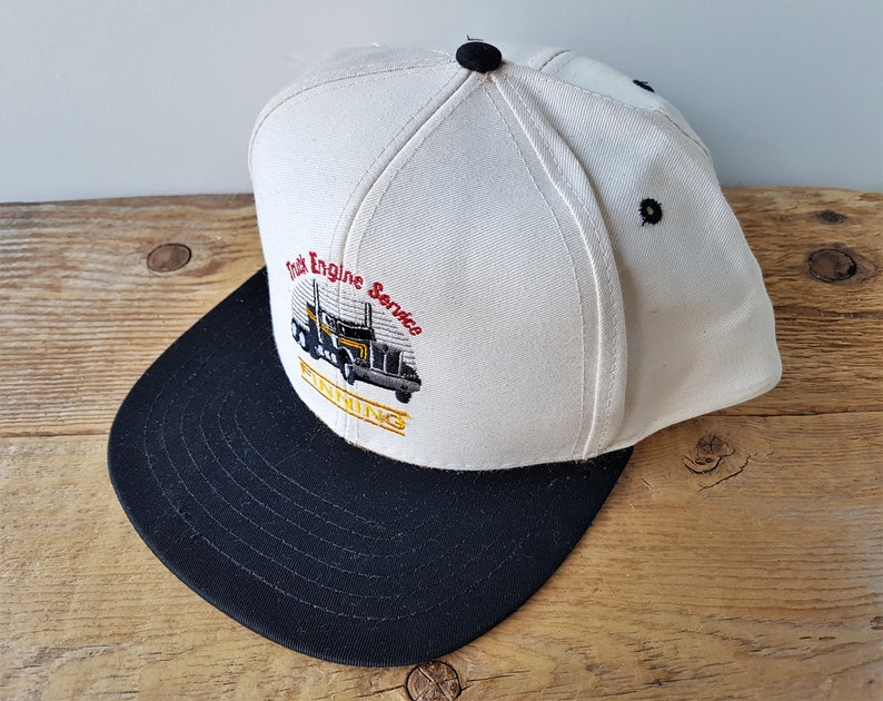 Op de afbeelding: Een wit en zwarte baseball cap met de tekst "Truck Engine Service FINNING" en een afbeelding van een vrachtwagen. De cap heeft een zwarte klep en een zwarte knop bovenop.