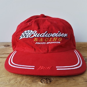 Op de afbeelding: Rode baseballcap met wit geborduurde tekst en accenten. De cap heeft het Budweiser Racing logo en het woord "Bud" op de klep. Trots sponsor van de racesport.