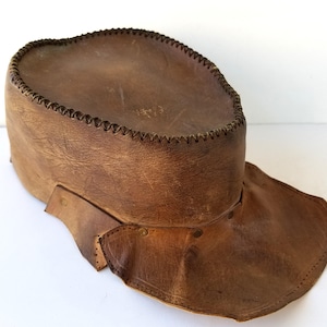 Puede incluir: Un sombrero de cuero marrón con ala ancha y corona cilíndrica. El sombrero presenta un borde cosido y está construido con costuras y remaches visibles. El cuero tiene un aspecto desgastado y texturizado.