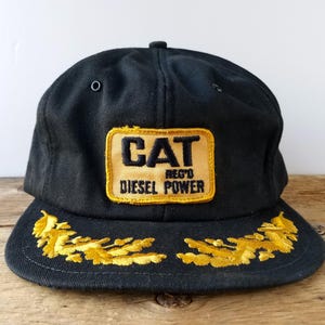 Può includere: Un cappellino da baseball nero con una toppa rettangolare gialla sul davanti che riporta il testo nero CAT REG'D DIESEL POWER. La visiera è decorata con motivi a foglie di quercia gialle ricamati su entrambi i lati della parte anteriore.
