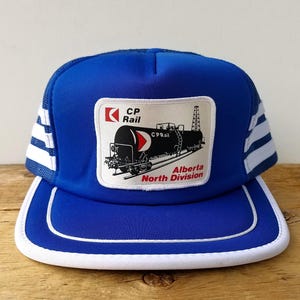 Könnte beinhalten: Blaue Trucker-Mütze mit weißem Schirm und weiß gestreiften Mesh-Seiten. Die Mütze hat einen rechteckigen Aufnäher mit dem CP Rail-Logo und einer Zugillustration sowie dem Text "Alberta North Division."