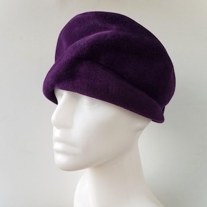 Puede incluir: Un sombrero tipo boina de color púrpura oscuro. El sombrero está hecho de un material suave y texturizado, con una corona redondeada y un ala doblada. El sombrero se exhibe en una cabeza de maniquí blanca, mostrando su forma y diseño.