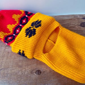 Puede incluir: Un pasamontañas tejido en colores brillantes. La parte superior es roja con un pompón negro y un patrón amarillo y negro. La abertura facial y el cuello son amarillos. El pasamontañas está hecho de un material suave y cálido.