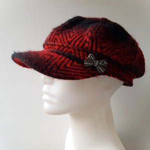 Puede incluir: Gorra de estilo newsboy con estampado de espiga rojo y negro y una pequeña visera. La gorra está adornada con un lazo plateado adornado con pedrería. El sombrero se muestra en una cabeza de maniquí blanca.