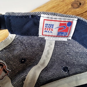 May include: A navy blue baseball cap with a white and blue checkered pattern on the inside. The cap has a curved brim and an adjustable strap. The label reads "ONE SIZE FITS ALL SPORTS SPECIALTIES".