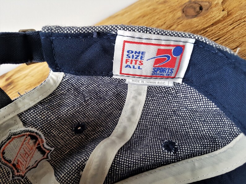 May include: A navy blue baseball cap with a white and blue checkered pattern on the inside. The cap has a curved brim and an adjustable strap. The label reads "ONE SIZE FITS ALL SPORTS SPECIALTIES".