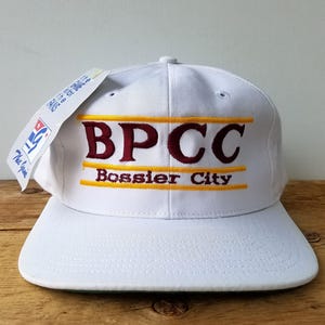 May include: White baseball cap with the text "BPCC Bossier City" embroidered in maroon and yellow. The cap has a flat brim and a tag attached to the side. The cap is sitting on a wooden surface.