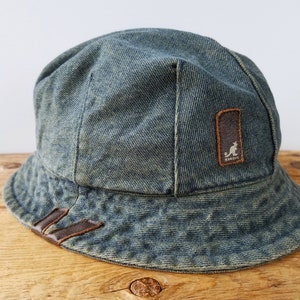 May include: A blue denim bucket hat with a brown leather patch featuring a kangaroo logo and the word "Kangol" in white lettering.
