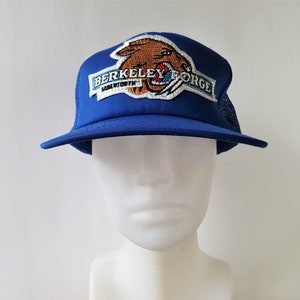 Może przedstawiać: Czapka truckerowa w kolorze niebieskim i białym z haftowanym logo w kolorze brązowym i białym, na którym widnieje napis "Berkeley Forge" i "Sabetha, Kansas".