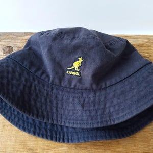K&ouml;nnte beinhalten: Marineblauer Fischerhut mit gelbem K&auml;nguru-Logo und dem Wort "KANGOL". Der Hut ist aus einem weichen Stoff gefertigt und hat eine breite Krempe. Ein klassisches Accessoire f&uuml;r Sonnenschutz und Freizeitkleidung.