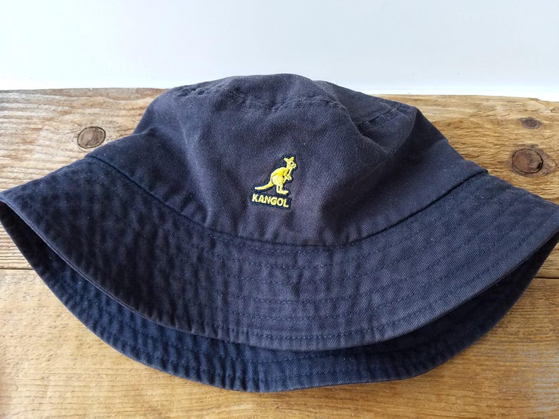 K&ouml;nnte beinhalten: Marineblauer Fischerhut mit gelbem K&auml;nguru-Logo und dem Wort "KANGOL". Der Hut ist aus einem weichen Stoff gefertigt und hat eine breite Krempe. Ein klassisches Accessoire f&uuml;r Sonnenschutz und Freizeitkleidung.