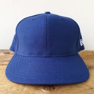May include: A solid blue baseball cap with a curved brim. The cap has a structured crown and a small white logo on the side. The hat is made of a durable fabric and is designed to be worn outdoors.