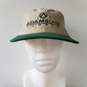 Puede incluir: Gorra de béisbol beige con visera verde. La gorra presenta el texto bordado "ADAMSLAKE LUMBER" en verde, junto con un logotipo verde. Se muestra sobre una cabeza de maniquí blanca.