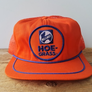 Op de afbeelding: Oranje truckerpet met een mesh achterkant en een platte klep. De pet heeft een cirkelvormige patch met de tekst "HOE-GRASS" en een logo, omlijnd in blauw. Gemaakt van stof en mesh, met een blauwe rand.