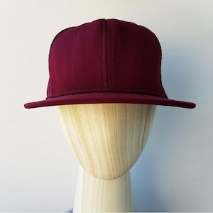May include: A burgundy baseball cap with a flat peak. The cap is displayed on a wooden mannequin head against a white background. The cap has a structured crown and a slightly textured fabric.