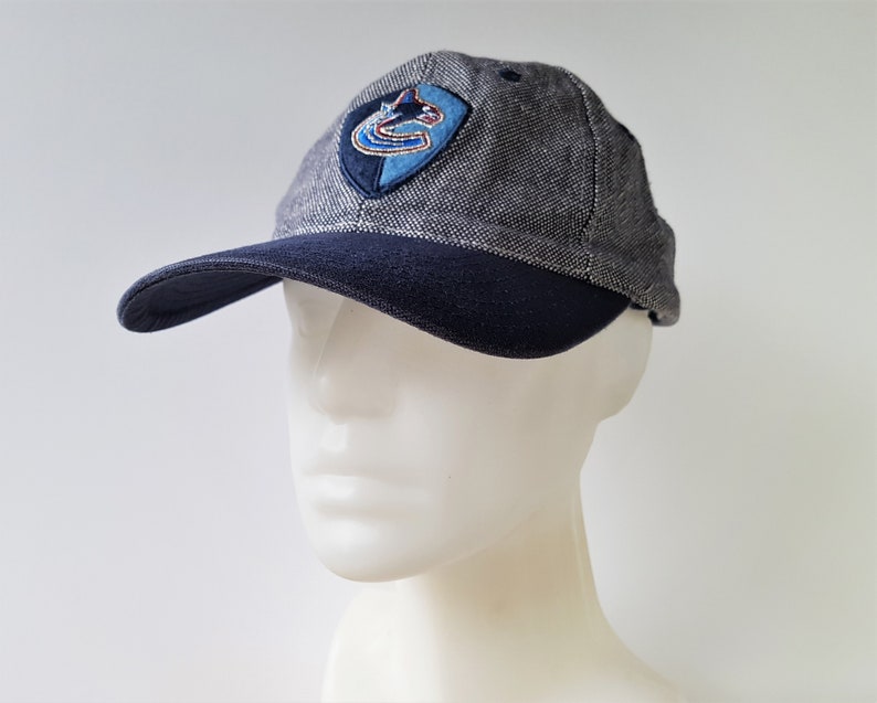 May include: A gray and navy blue baseball cap with a curved brim. The cap features a team logo patch on the front. The cap is made of a textured fabric and has a classic six-panel design.