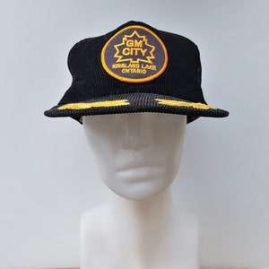 May include: Black corduroy baseball cap with a yellow and black embroidered patch that reads "GM CITY KIRKLAND LAKE ONTARIO"