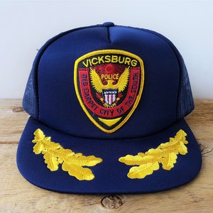 May include: A navy blue trucker hat with a yellow and red embroidered patch that says "Vicksburg Police City of the South" and two gold embroidered oak leaf accents.