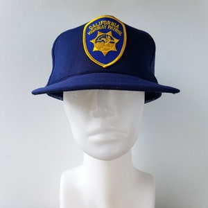 May include: A navy blue trucker hat with a mesh back and a curved brim. The hat features a patch with the words "CALIFORNIA HIGHWAY PATROL" and a gold star emblem on a blue background. The hat is displayed on a white mannequin head.
