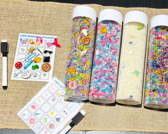 May include: Four clear plastic bottles filled with different colored rice and small objects. The bottles are labeled "I Spy With My Little Eye".