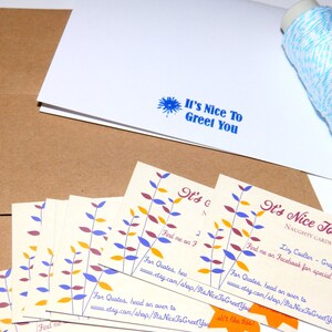 May include: A collection of greeting cards with the text "It's Nice To Greet You" in blue. The cards have a floral design in yellow, purple, and orange. Kraft paper envelopes and a spool of blue and white twine are also visible.