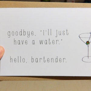 May include: A white greeting card with the text "goodbye, 'I'll just have a water.' hello, bartender." A simple drawing of a martini glass with an olive is on the right side. The card is held by a hand.