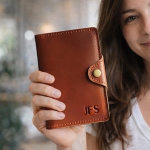 May include: A brown leather passport holder with the initials "JFS" and a navy blue United States passport. The passport holder has a button closure. The passport has the word "PASSPORT" on the cover.