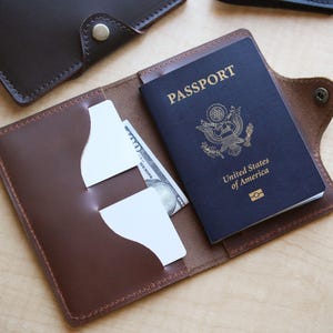 May include: Open brown leather passport holder with a dark blue passport and a US $100 bill inside. The passport is embossed with "PASSPORT" and "United States of America". The holder has card slots and a snap closure.
