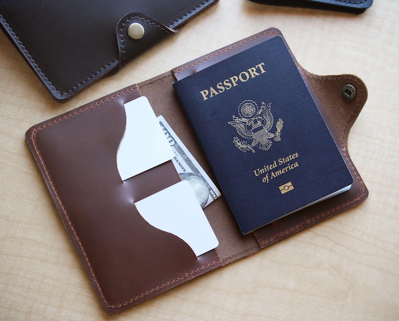May include: Open brown leather passport holder with a dark blue passport and a US $100 bill inside. The passport is embossed with "PASSPORT" and "United States of America". The holder has card slots and a snap closure.