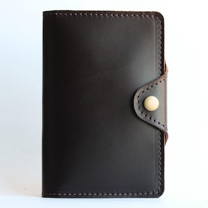 May include: Dark brown leather wallet with a button closure. The wallet is rectangular with rounded corners and features a light-colored button. The edges are stitched with a lighter brown thread. The background is white.