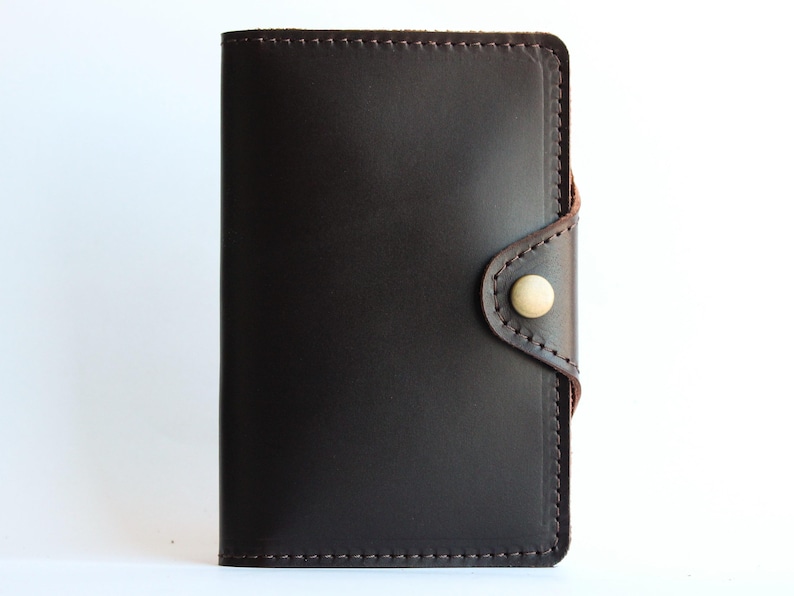 May include: Dark brown leather wallet with a button closure. The wallet is rectangular with rounded corners and features a light-colored button. The edges are stitched with a lighter brown thread. The background is white.
