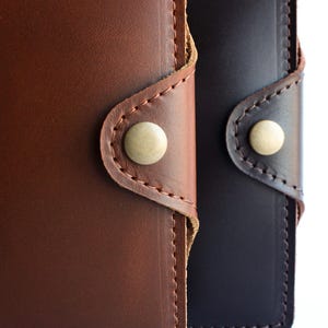 May include: Close-up of two leather wallets, one in rich brown and the other in dark navy. Each wallet features a button closure with a light-colored button and detailed stitching.
