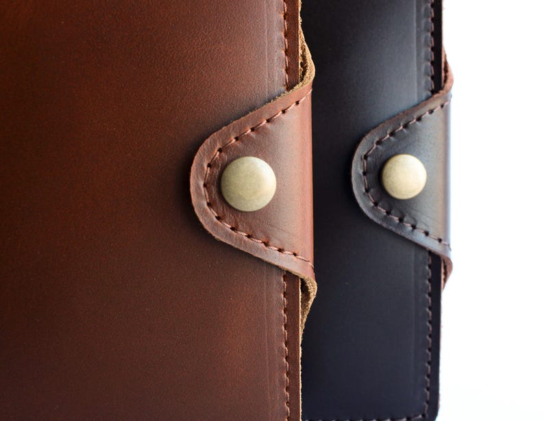 May include: Close-up of two leather wallets, one in rich brown and the other in dark navy. Each wallet features a button closure with a light-colored button and detailed stitching.
