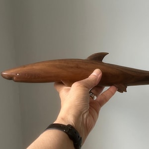 May include: A wooden shark sculpture, with a smooth, streamlined design. The shark is carved from dark brown wood, with fins and a tail. The sculpture is held in a hand, showing its size and detail. The shark is approximately 40 cm long.