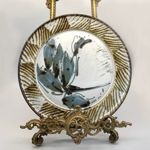 May include: A ceramic plate with a brown and white striped border and a blue and brown floral design. The plate is displayed on a gold metal stand.