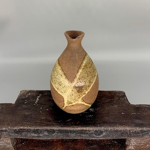 May include: A small, brown ceramic vase with a light brown and yellow glaze. The vase has a rounded body and a narrow neck.